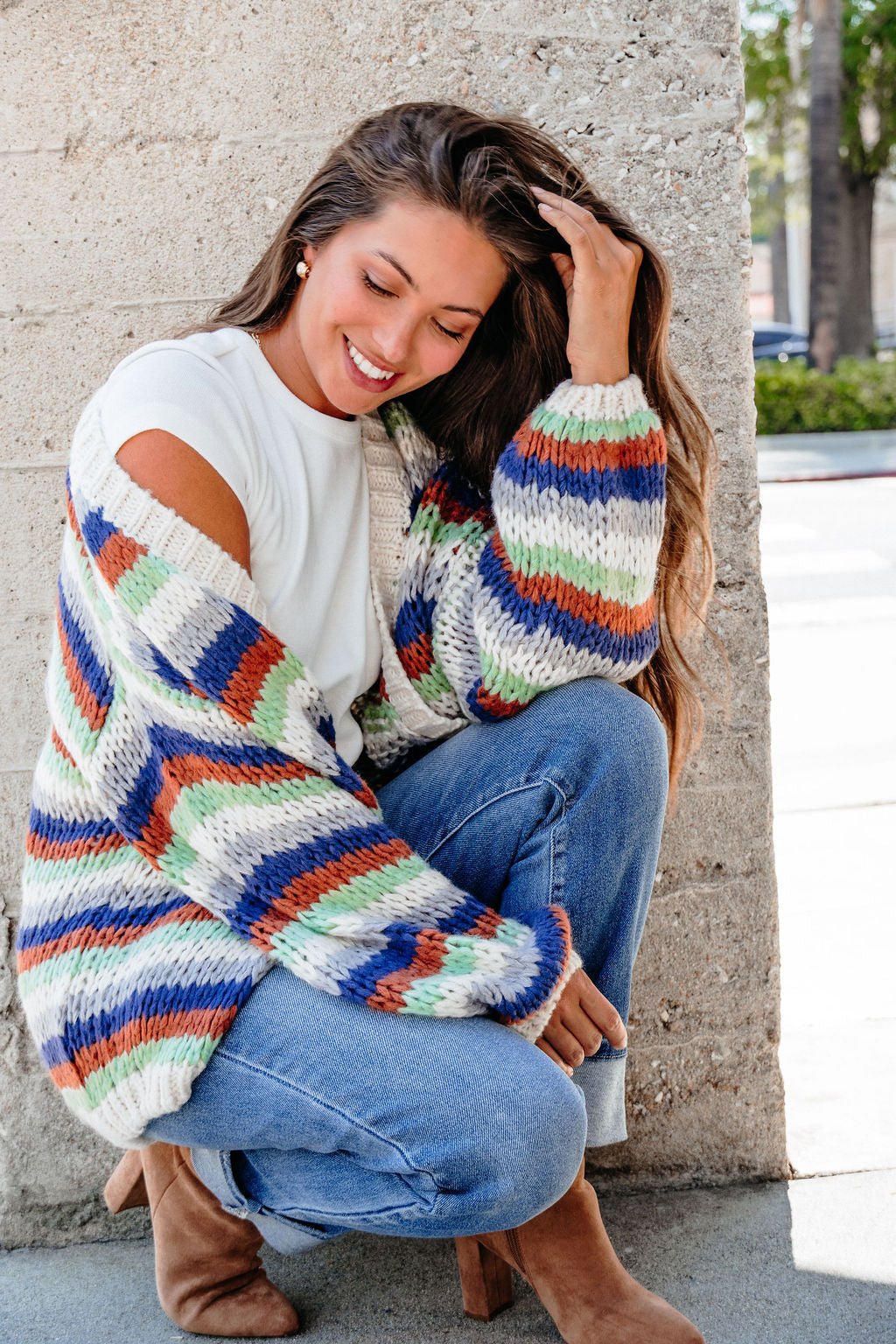 Smiling woman squats by a wall, wearing the Multi Striped Open Front Sweater Cardigan - FINAL SALE, jeans, and brown boots.