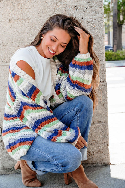 Smiling woman squats by a wall, wearing the Multi Striped Open Front Sweater Cardigan - FINAL SALE, jeans, and brown boots.