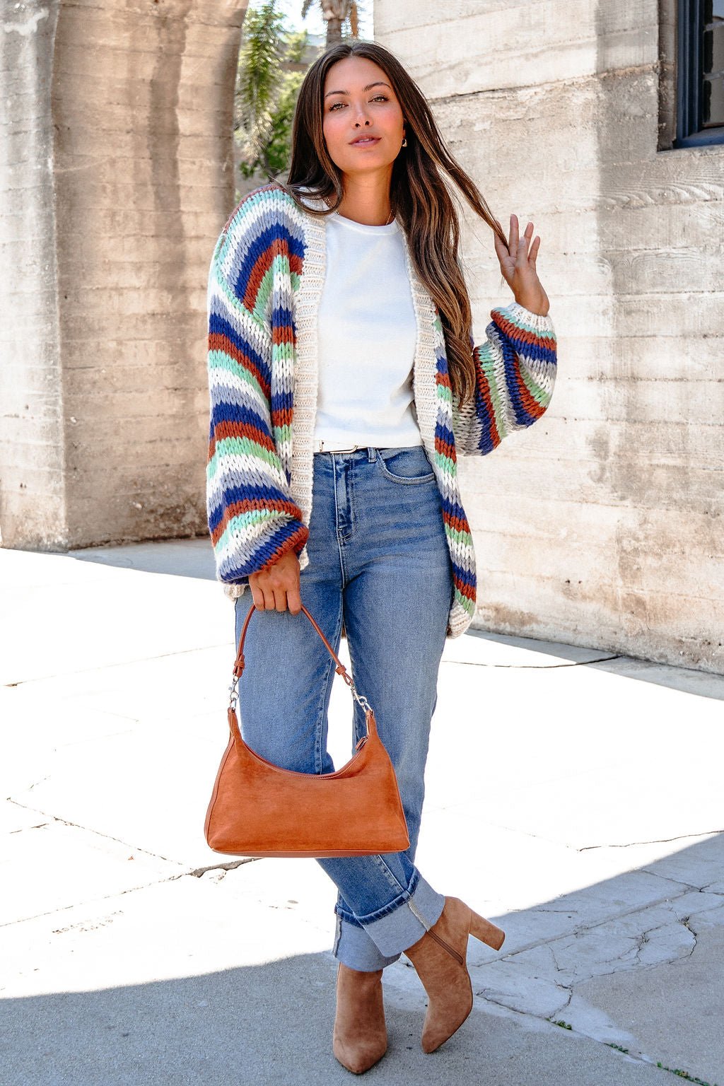 A woman wears jeans, a white top, and the Multi Striped Open Front Sweater Cardigan while posing outdoors with a tan handbag.