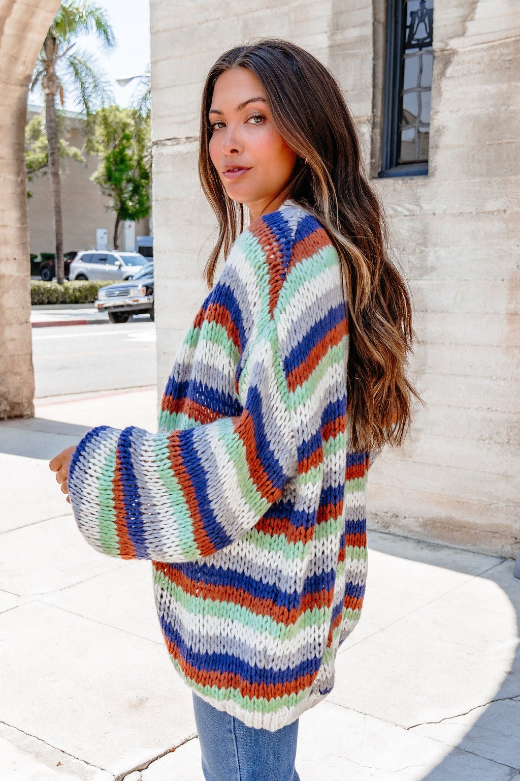 A woman with long hair wears the Multi Striped Open Front Sweater Cardigan, standing outdoors by a sunlit stone building.
