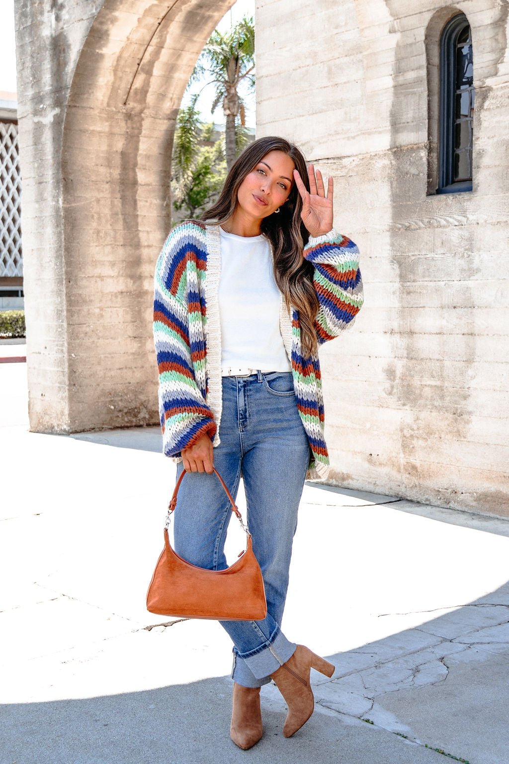 Woman in cuffed jeans, white shirt, and Multi Striped Open Front Sweater Cardigan stands outdoors waving, holding a tan handbag.