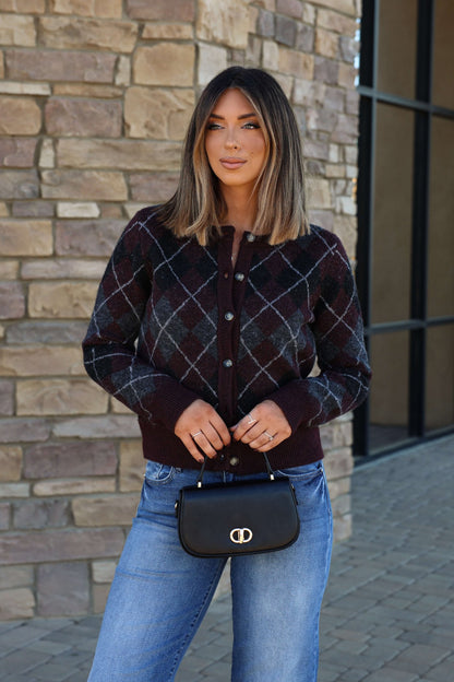 Woman with straight, shoulder-length hair in an argyle sweater and jeans holds the Nadine Black Vegan Leather Crossbody Bag by a stone wall.