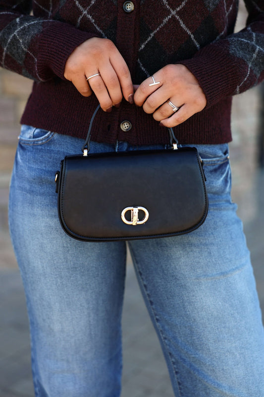 A person in blue jeans and a maroon sweater holds the Nadine Black Vegan Leather Crossbody Bag - DOORBUSTER.