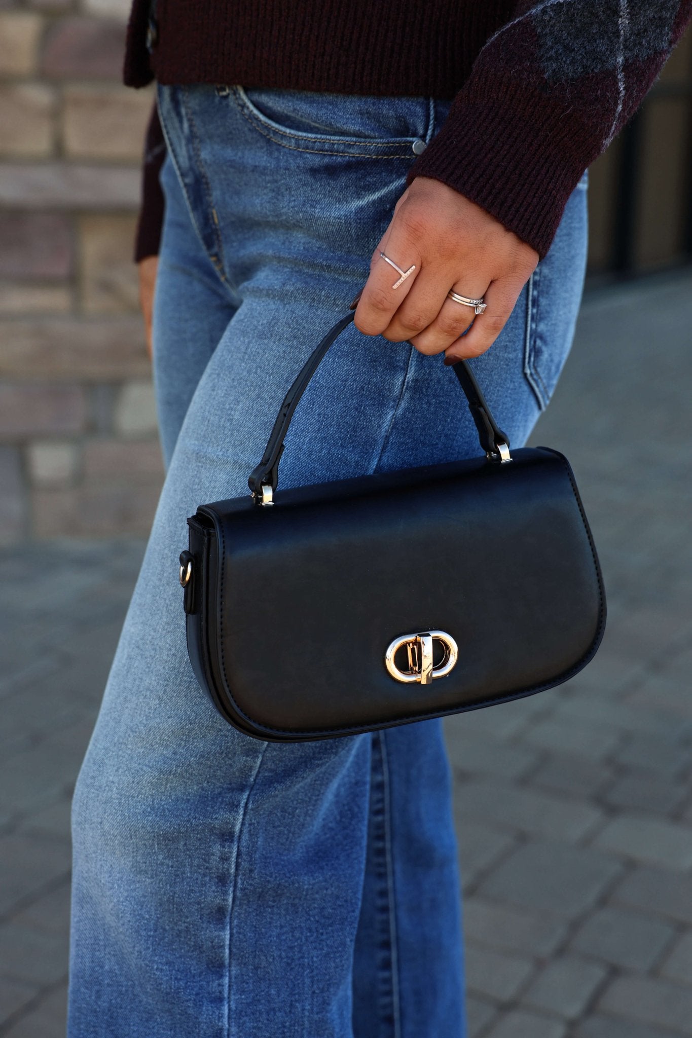 A person in blue jeans holds the Nadine Black Vegan Leather Crossbody Bag - DOORBUSTER, wearing silver rings.
