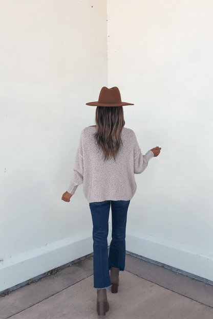 Woman stands in a corner facing the wall, wearing a Natasha Chunky Cable Knit Sweater in grey, brown hat, jeans, and boots.