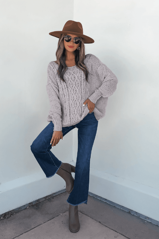 Woman in Natasha Grey Cable Knit Sweater, tan hat, sunglasses, jeans, and taupe boots poses by a white wall.
