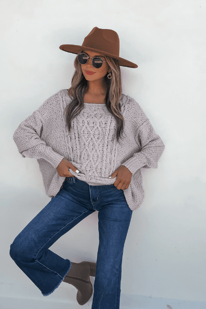 Woman wears sunglasses, a tan hat, blue jeans, ankle boots, and the Natasha Chunky Cable Knit Sweater in grey against a white wall.
