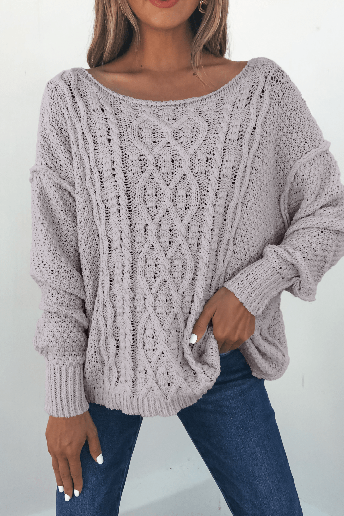 Woman wearing the Natasha Chunky Cable Knit Sweater in grey, paired with blue jeans and holding the cozy sweater hem.