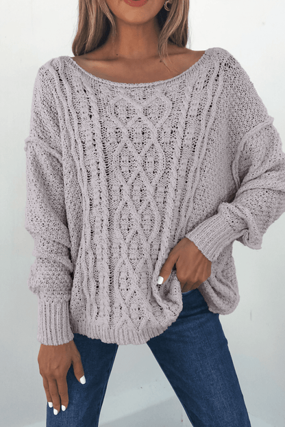 Woman wearing the Natasha Chunky Cable Knit Sweater in grey, paired with blue jeans and holding the cozy sweater hem.