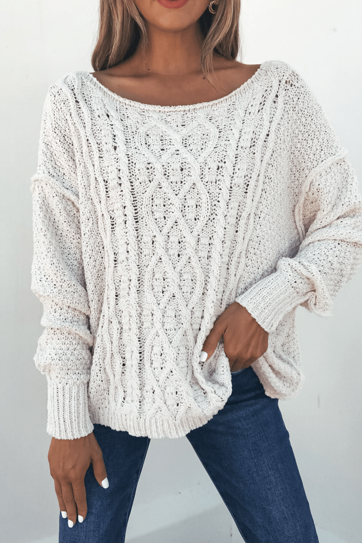 Woman wears the Natasha Ivory Cable Knit Sweater and blue jeans, posing with one hand on her hip against a plain light background.
