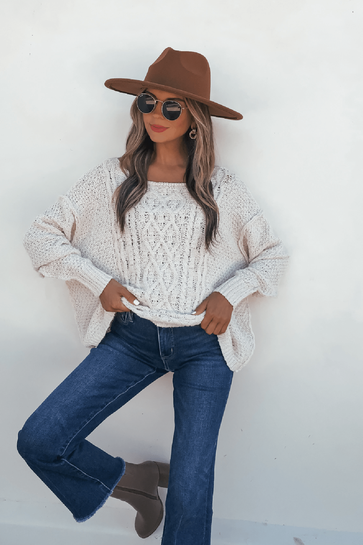 Woman in a brown hat, sunglasses, Natasha Ivory Cable Knit Sweater, blue jeans, and taupe boots poses by a white wall.