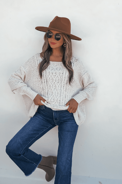 Woman in a brown hat, sunglasses, Natasha Ivory Cable Knit Sweater, blue jeans, and taupe boots poses by a white wall.