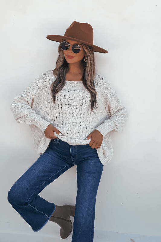 Woman in a brown hat, sunglasses, Natasha Ivory Cable Knit Sweater, blue jeans, and taupe boots poses by a white wall.