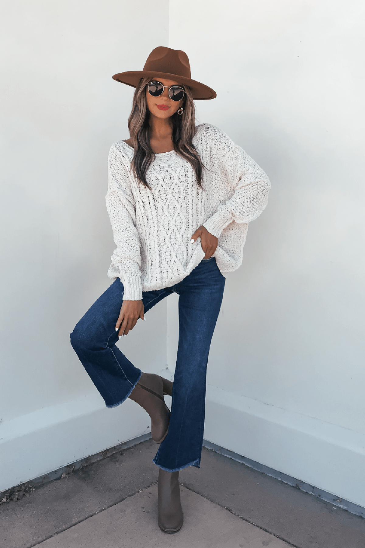 Woman in wide-brim hat, sunglasses, and blue jeans poses in the Natasha Ivory Cable Knit Sweater and taupe boots.