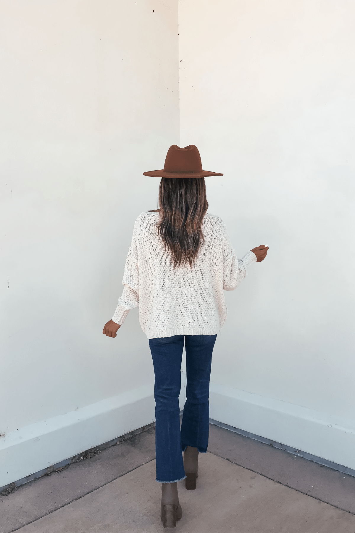 Woman stands in a corner, facing away, wearing the Natasha Ivory Cable Knit Sweater with blue jeans and brown boots.