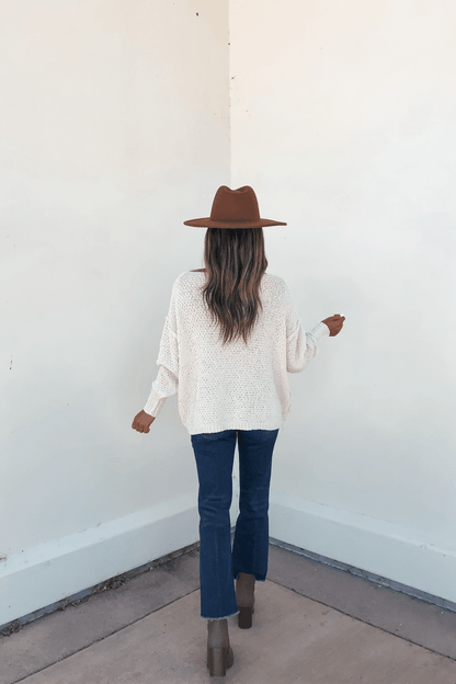 Woman stands in a corner, facing away, wearing the Natasha Ivory Cable Knit Sweater with blue jeans and brown boots.