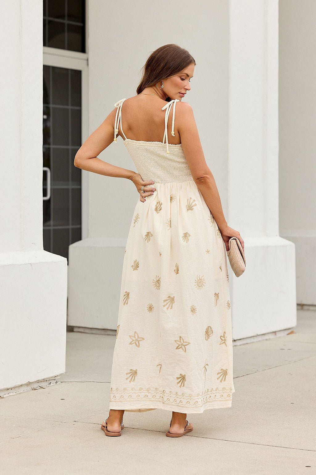 Woman in the Natural Beauty Embroidered Maxi Dress, cream with gold patterns and tie straps, holds a clutch near a white building.