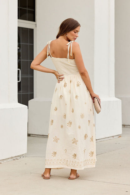 Woman in the Natural Beauty Embroidered Maxi Dress, cream with gold patterns and tie straps, holds a clutch near a white building.