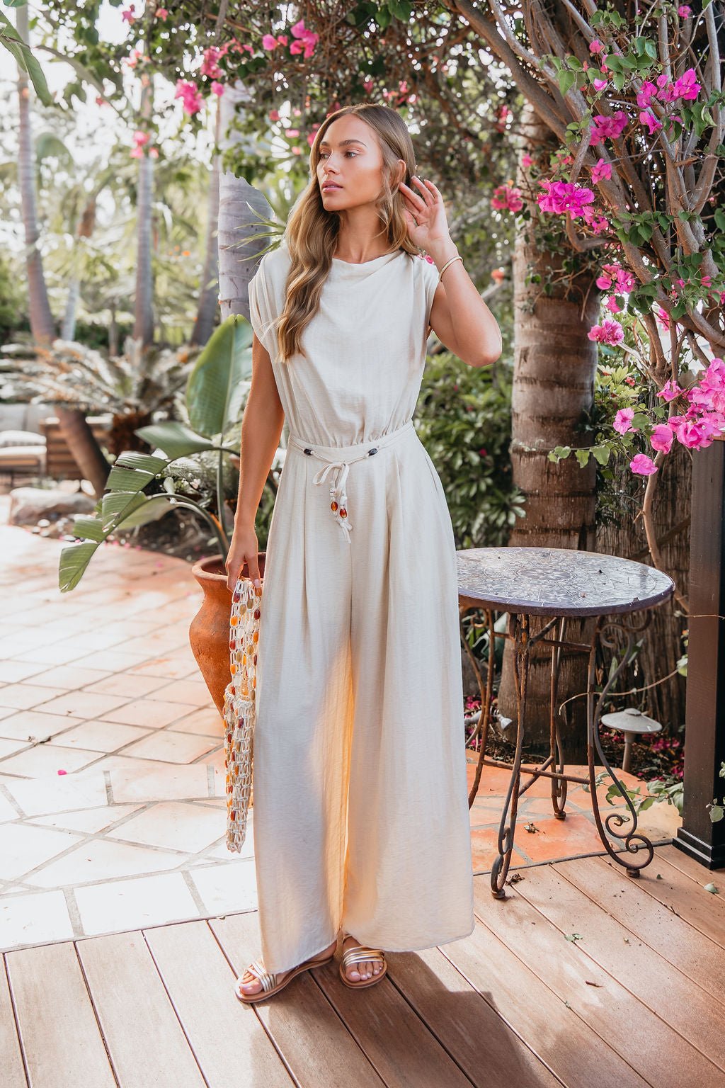 Woman in the Natural Belted Wide Leg Linen Jumpsuit stands on a patio with plants and pink flowers, holding a woven bag.