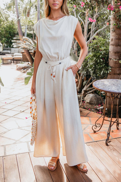 Woman models the Natural Belted Wide Leg Linen Jumpsuit - FINAL SALE, styled on a sunny patio with a beaded bag and plants.