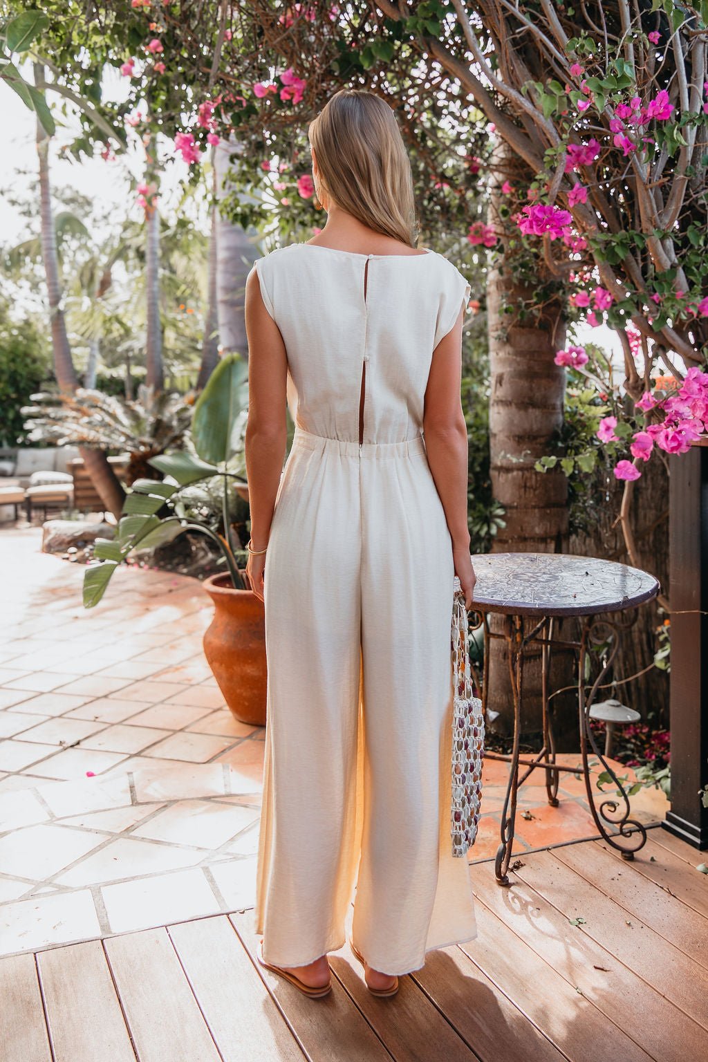 A woman wears the Natural Belted Wide Leg Linen Jumpsuit—FINAL SALE—standing on a patio near pink flowers and outdoor furniture.