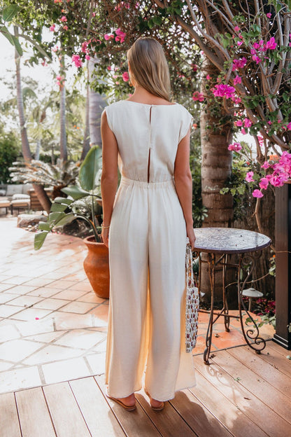A woman wears the Natural Belted Wide Leg Linen Jumpsuit—FINAL SALE—standing on a patio near pink flowers and outdoor furniture.