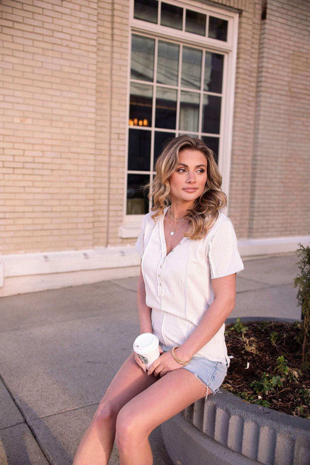 A woman with wavy blonde hair wears the Natural Contrast Trim Button Up Top, sitting on a planter edge holding coffee by a brick building.