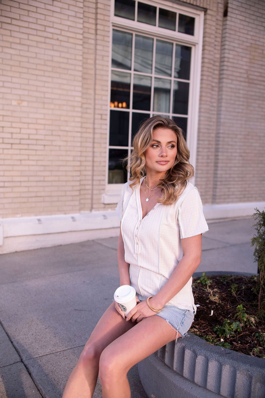 A woman in a Natural Contrast Trim Button Up Top and denim shorts sits on a planter edge with coffee in front of a brick building.