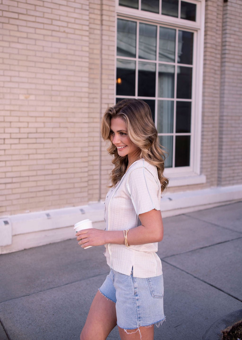 A woman in a Natural Contrast Trim Button Up Top and denim shorts walks outside, holding a coffee cup and smiling.