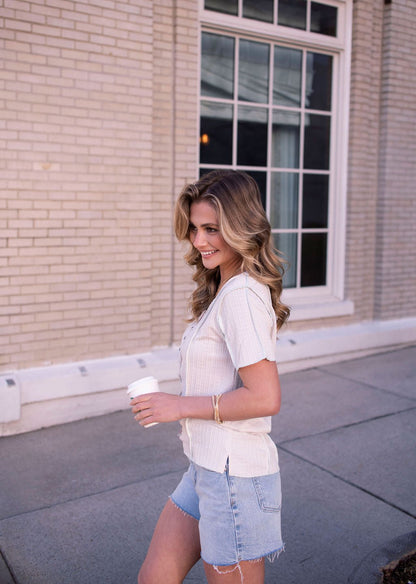 A woman in a Natural Contrast Trim Button Up Top and denim shorts walks outside, holding a coffee cup and smiling.