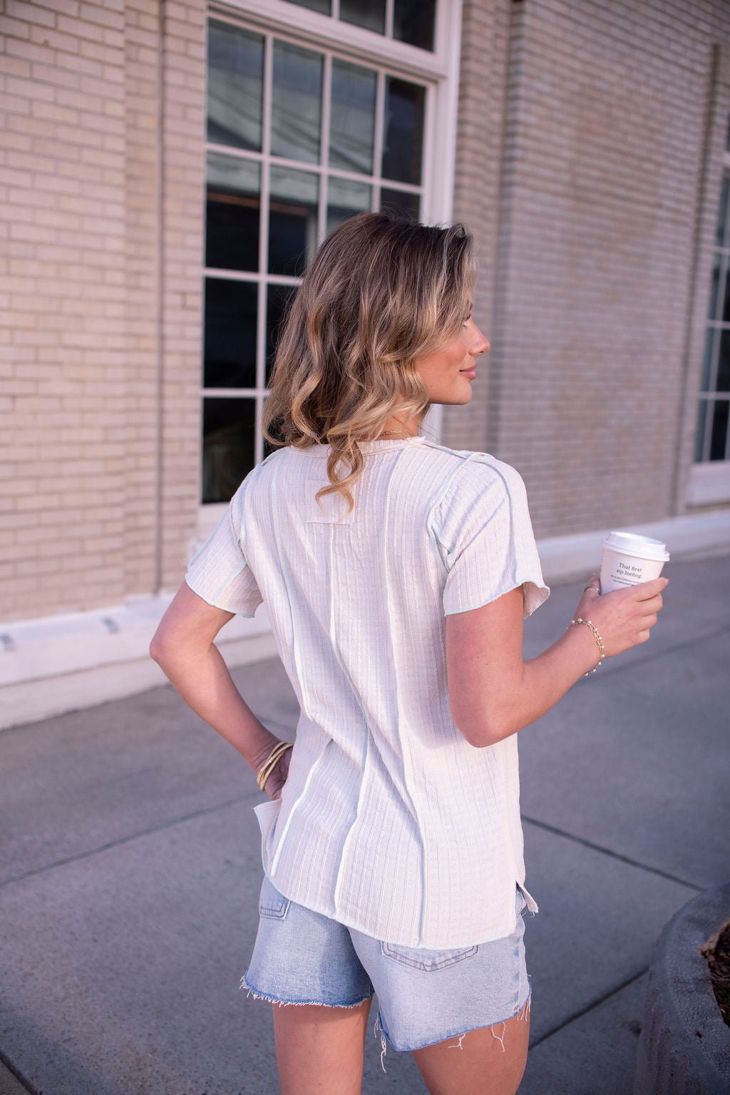 A woman with wavy blonde hair holds a coffee cup, wearing the Natural Contrast Trim Button Up Top and denim shorts outside.
