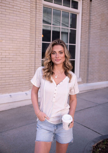 Woman with wavy blonde hair in a Natural Contrast Trim Button Up Top and denim shorts holds a coffee cup by a brick building.