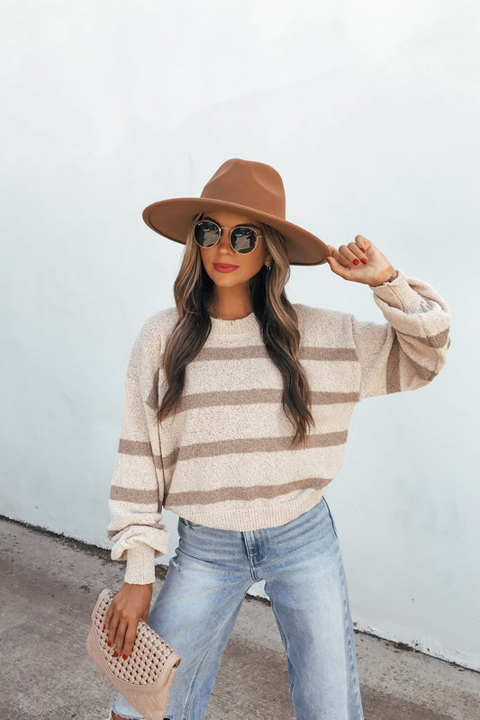 Woman in a Natural & Mocha Striped Sweater, jeans, hat, and sunglasses holds a beige clutch—an essential fall look.