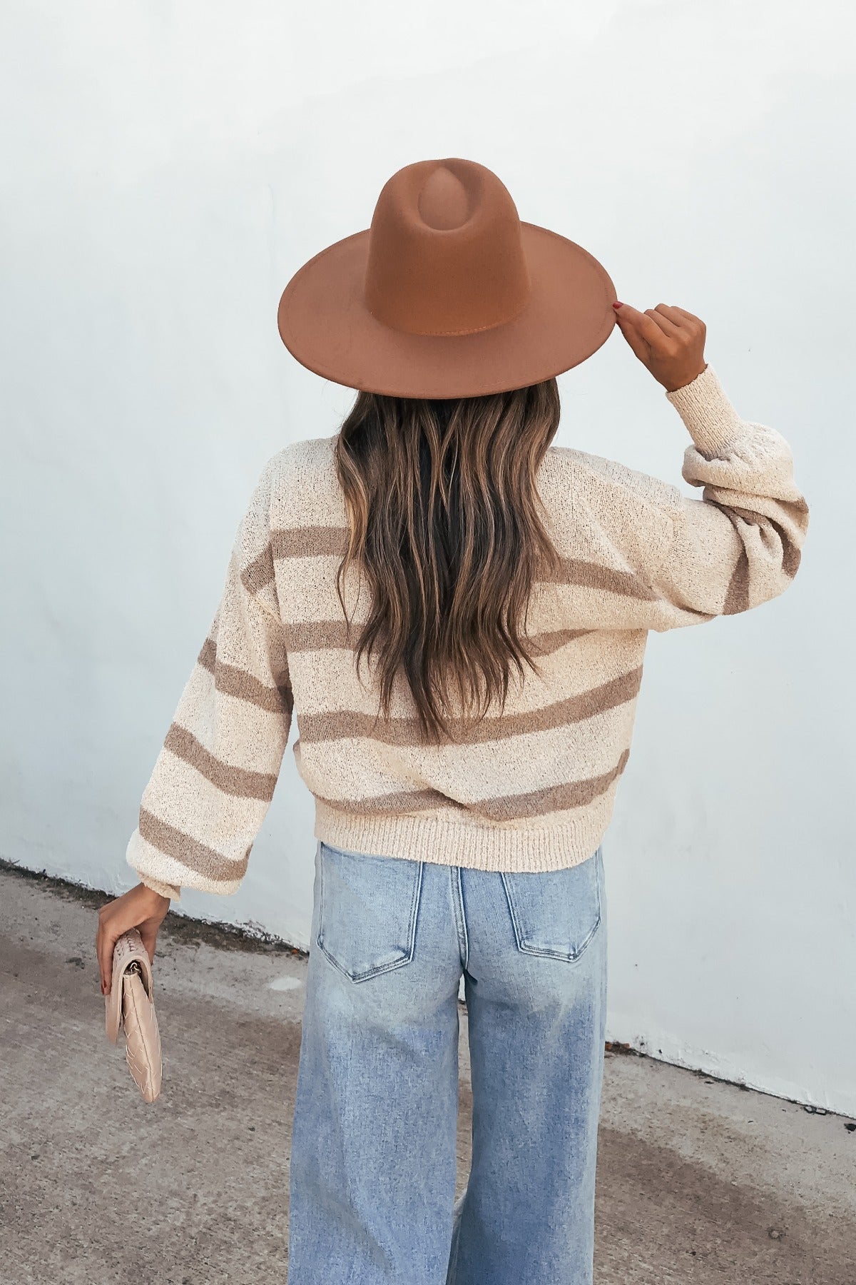 Individual in a wide-brimmed hat, light jeans, and the Natural & Mocha Striped Sweater faces a white wall holding a clutch.