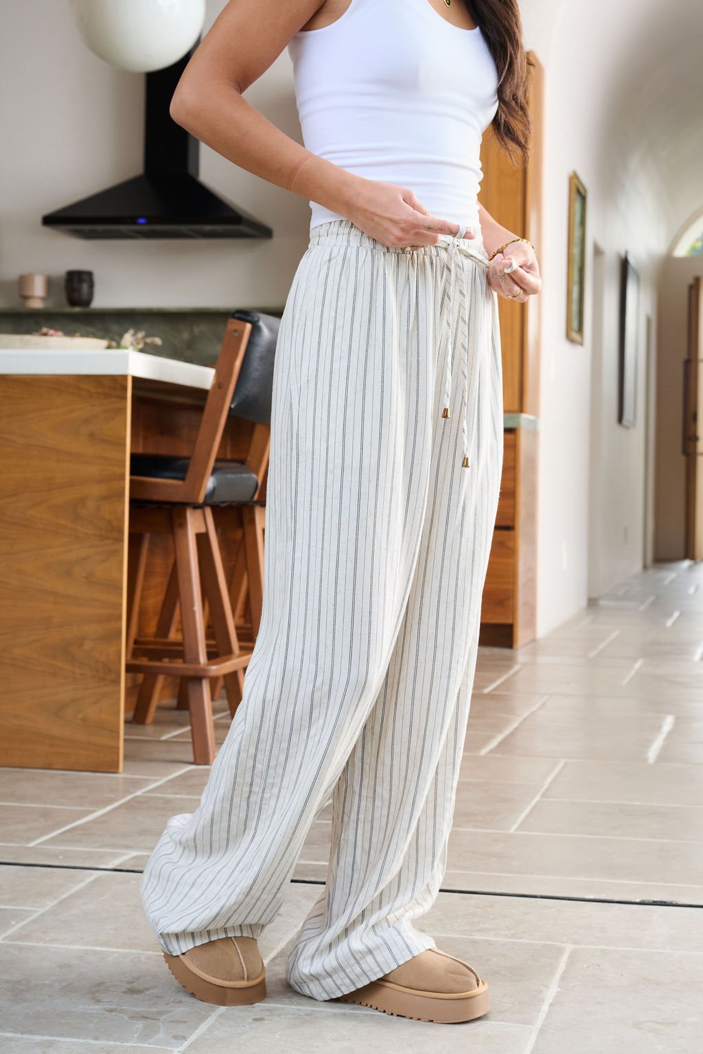 A woman indoors wears Natural Striped Linen Drawstring Pants, a white tank top, and tan slippers, holding her waistband.