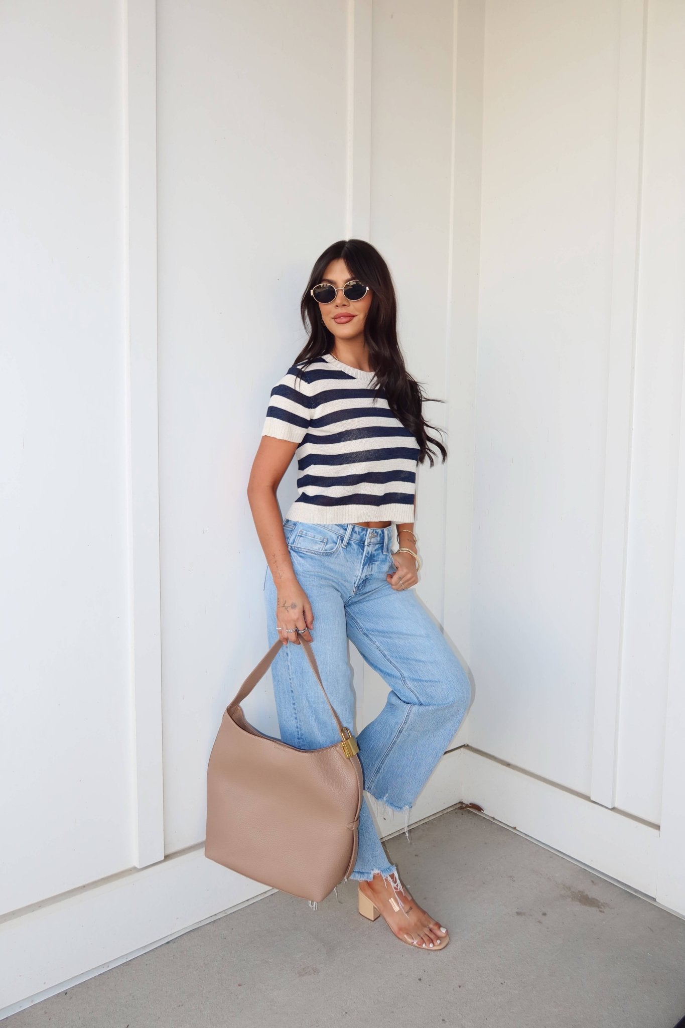 Woman wears a Navy and Cream Striped Short Sleeve Sweater with light blue jeans, sunglasses, and a taupe handbag against a white wall.