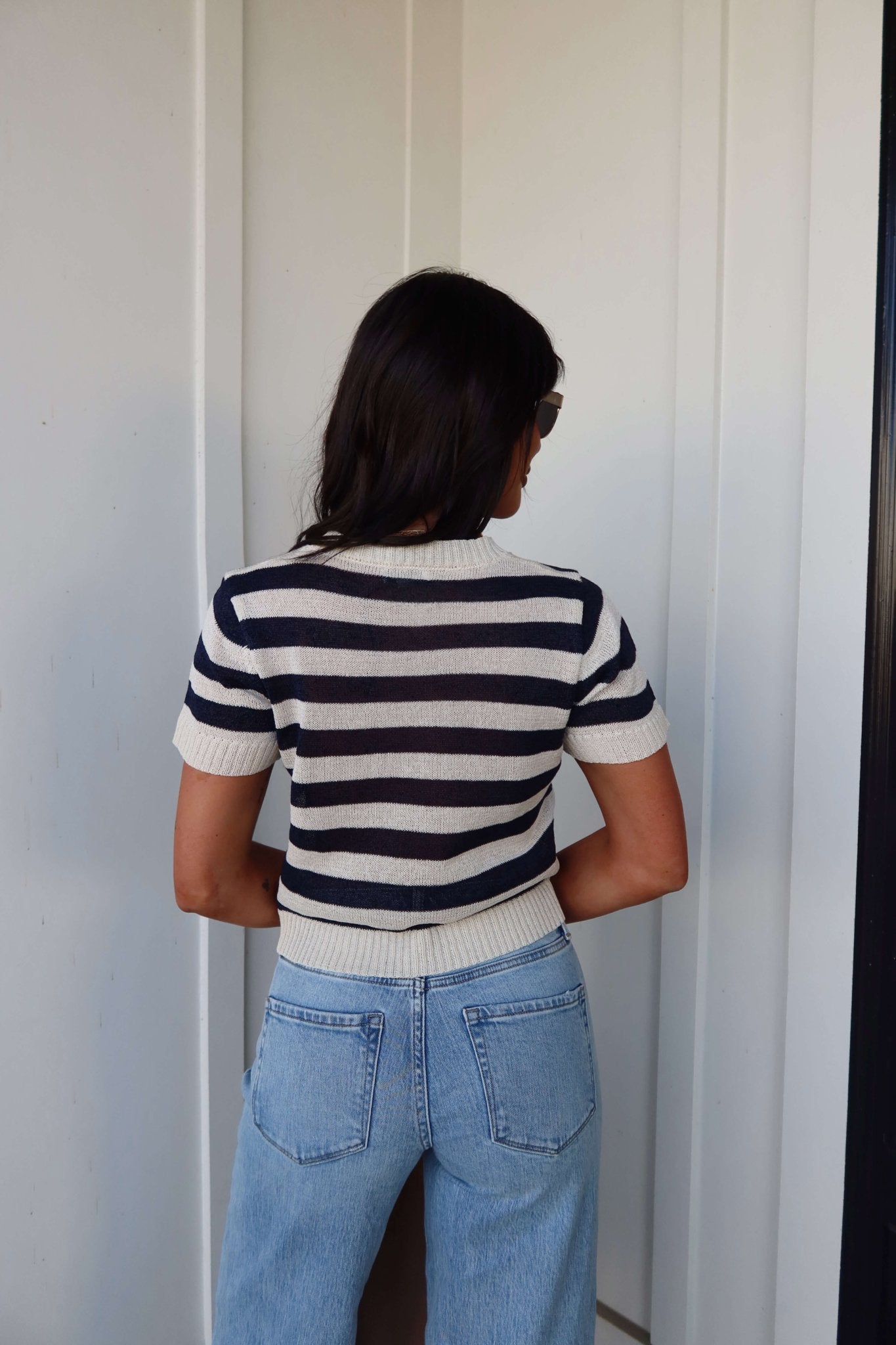 Woman with long dark hair wears a Navy and Cream Striped Short Sleeve Sweater and light blue jeans, back to the camera.