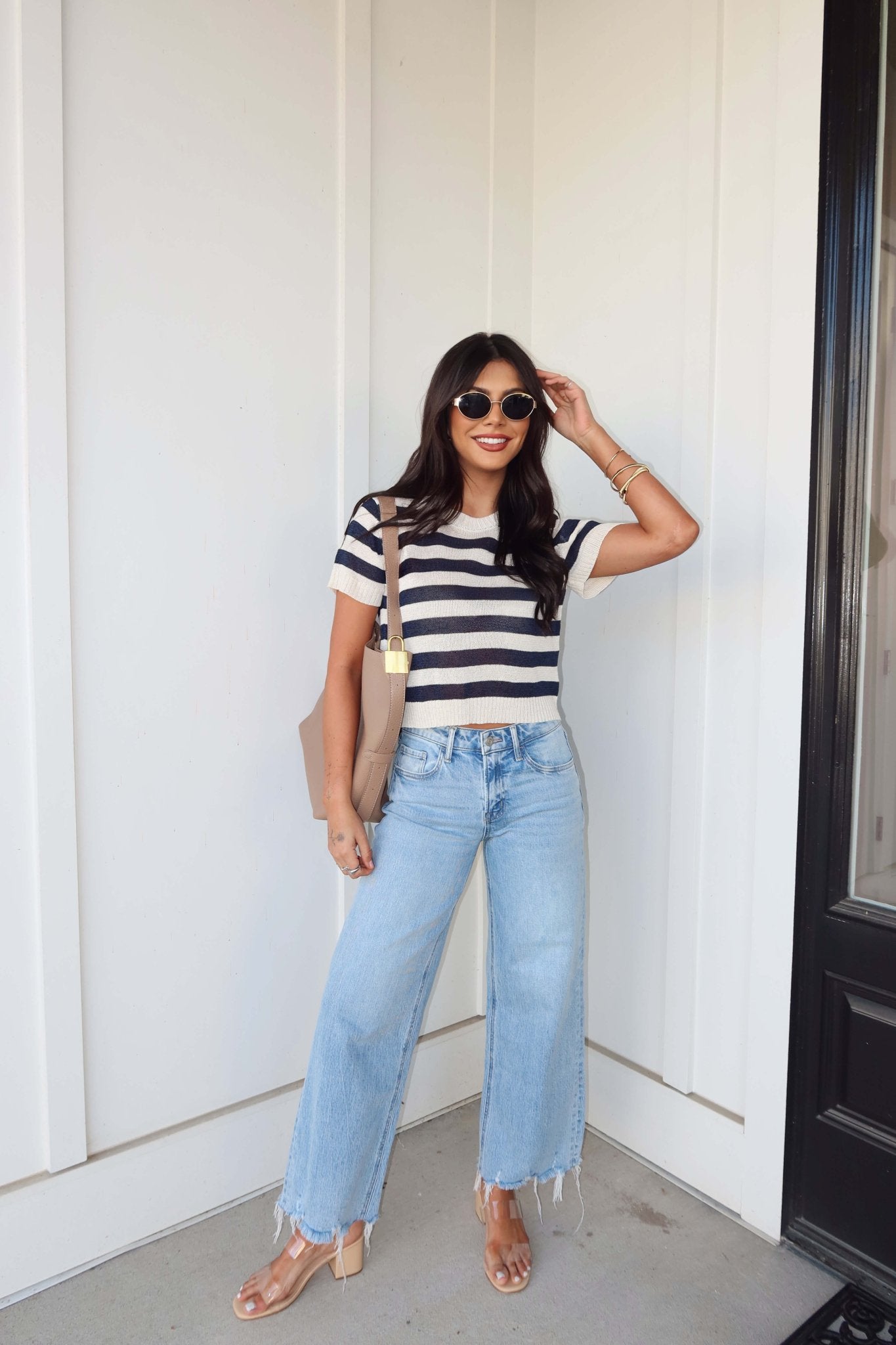Woman wearing a Navy and Cream Striped Short Sleeve Sweater, wide-leg jeans, sunglasses, and sandals by a white wall with black door.