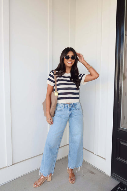 Woman wearing a Navy and Cream Striped Short Sleeve Sweater, wide-leg jeans, sunglasses, and sandals by a white wall with black door.