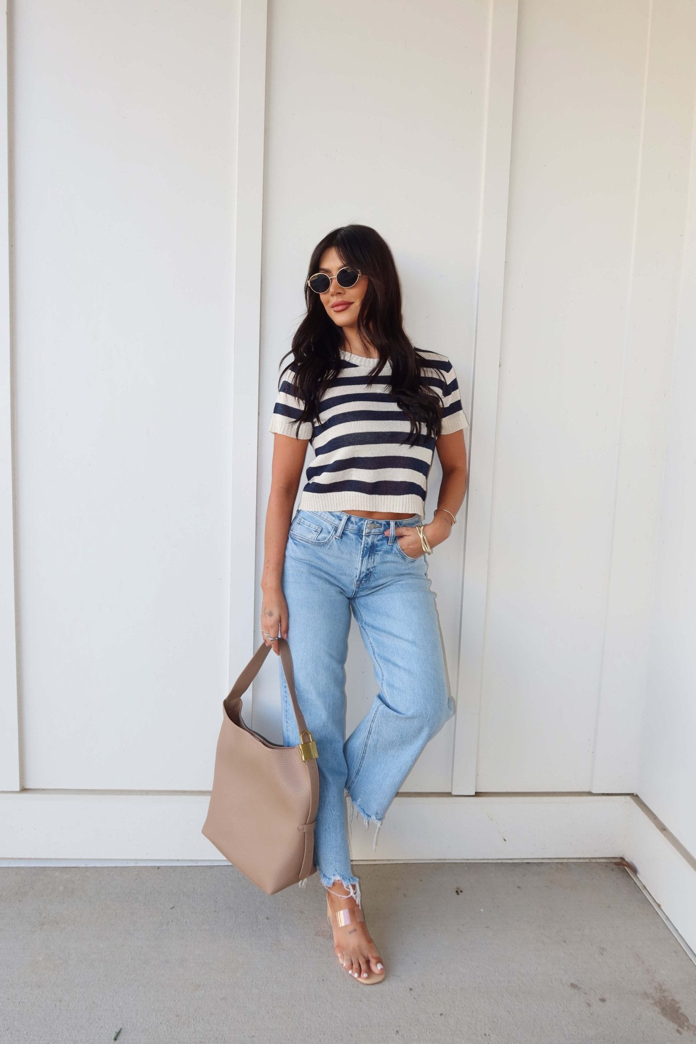 Woman in a Navy and Cream Striped Short Sleeve Sweater, light blue jeans, sandals, sunglasses, and beige handbag by a white wall.