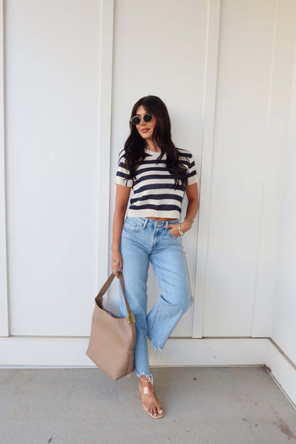 Woman in a Navy and Cream Striped Short Sleeve Sweater, light blue jeans, sandals, sunglasses, and beige handbag by a white wall.