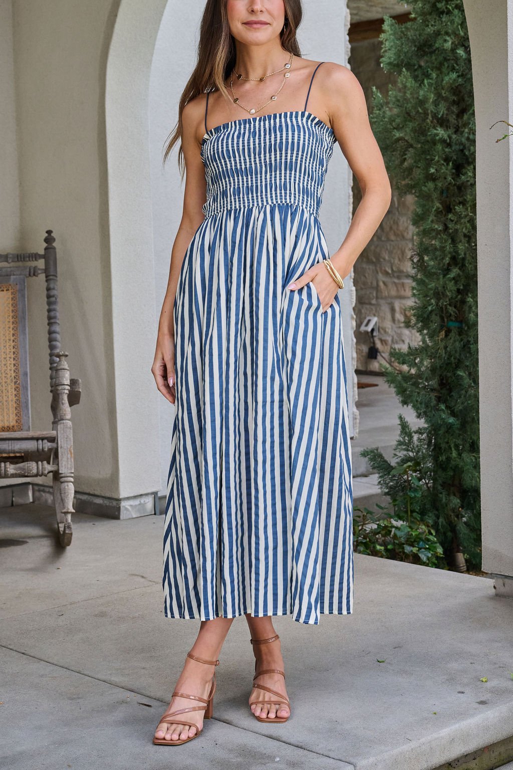 Woman wearing a Navy and Ivory Striped Midi Dress stands in sandals on a porch near greenery.