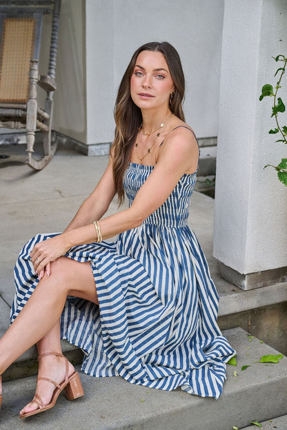 A woman in the Navy and Ivory Striped Midi Dress sits on steps, gazing at the camera with a relaxed expression.