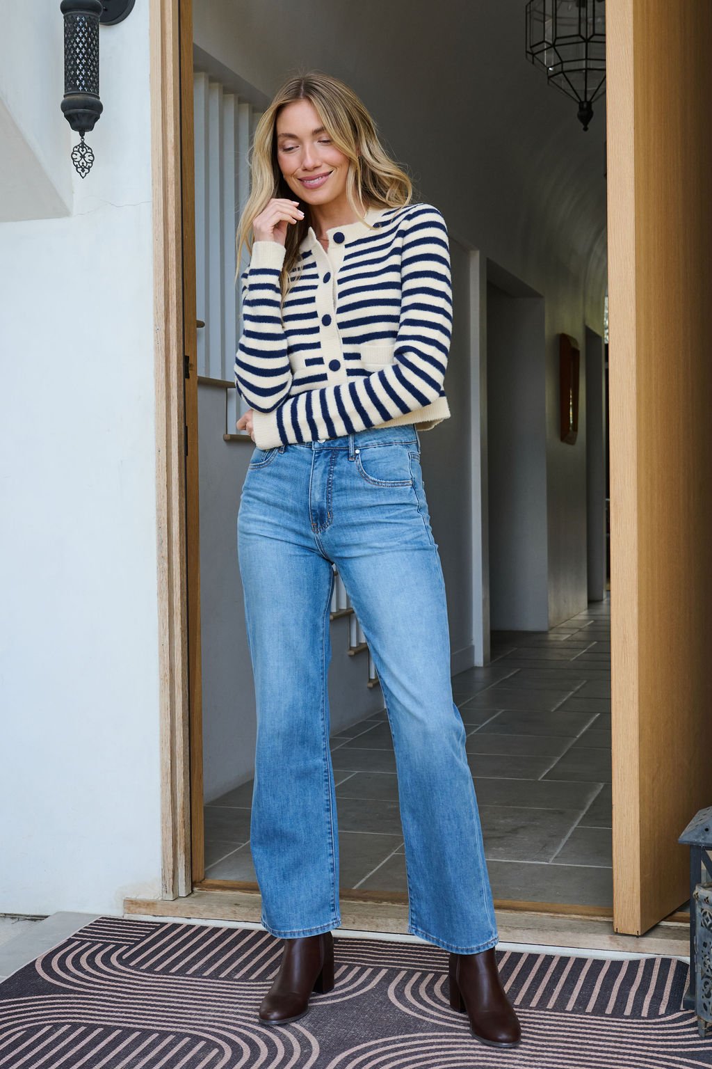 A woman in a Navy and Tan Striped Button Down Cardigan and blue jeans stands smiling in a doorway, hand resting on her chin.