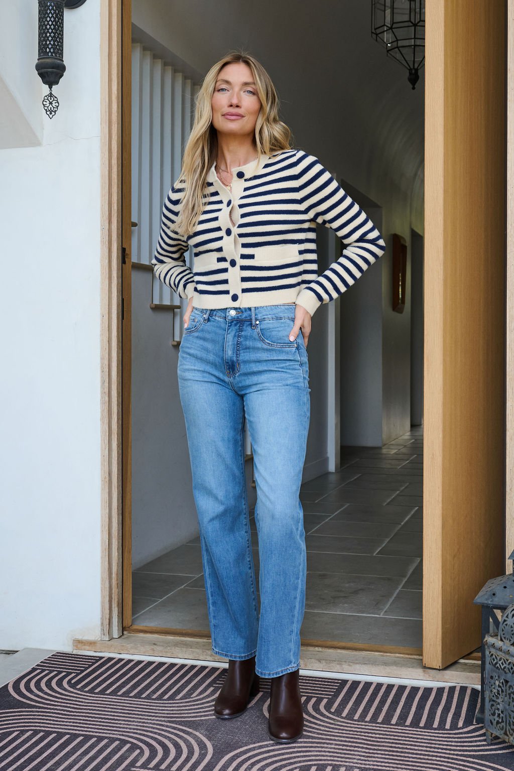 A woman stands in a doorway wearing a Navy and Tan Striped Button Down Cardigan, blue jeans, and brown boots, hands in pockets.