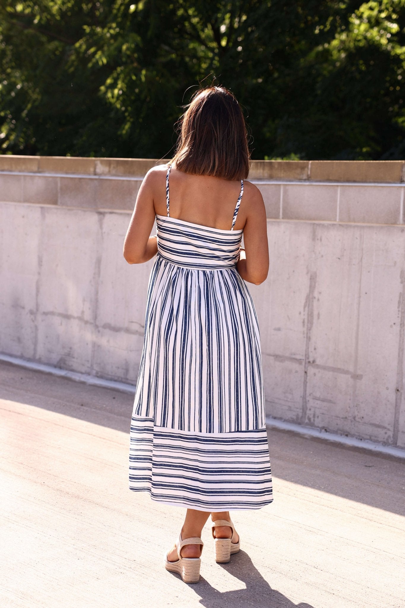 Woman with shoulder-length hair outdoors models a Navy and White Striped Tie Front Midi Dress by a concrete wall.