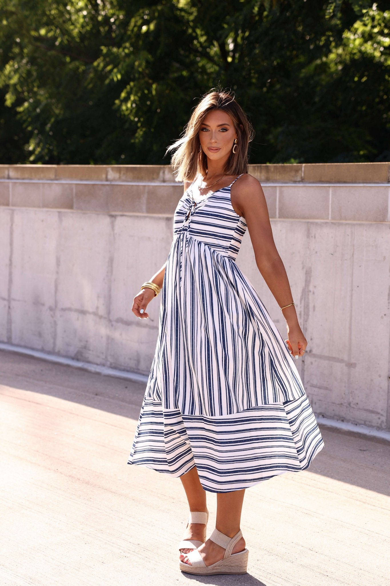 Woman outdoors in a Navy and White Striped Tie Front Midi Dress, holding her skirt amid sunlight and greenery.