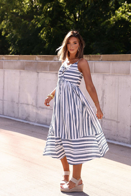 Woman outdoors in a Navy and White Striped Tie Front Midi Dress, holding her skirt amid sunlight and greenery.