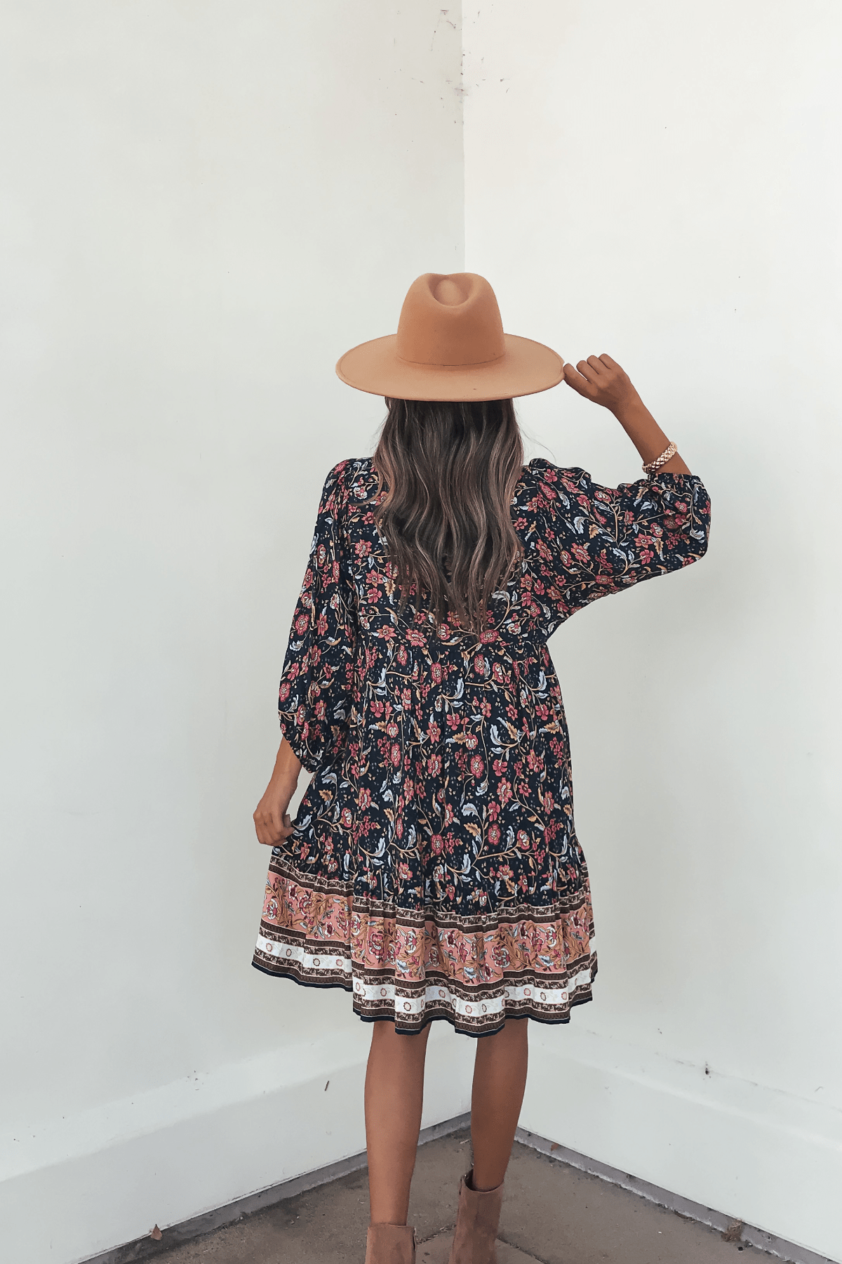 Woman in the Navy Floral Border Print Mini Dress - FINAL SALE, styled with a tan hat and boots, stands against a white wall.