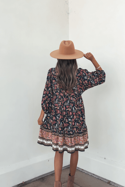 Woman in the Navy Floral Border Print Mini Dress - FINAL SALE, styled with a tan hat and boots, stands against a white wall.